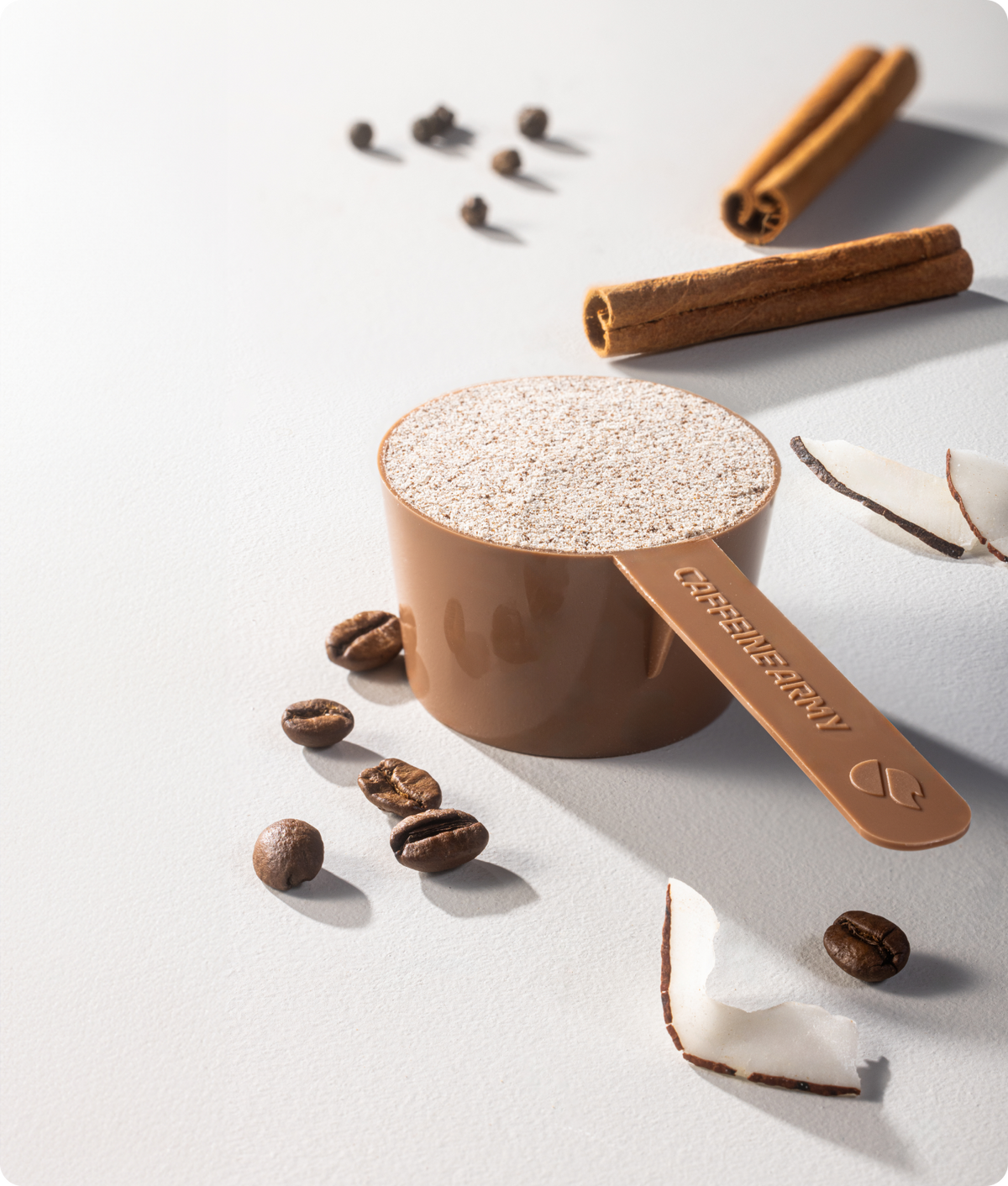 Colher dosadora cheia de SuperCoffee entre grãos de café, lascas de coco e paus de canela sobre fundo claro.