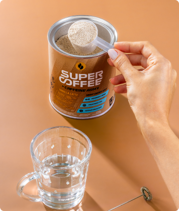 Pessoa retirando uma colher dosadora de SuperCoffee Vanilla Latte da lata, com copo de água e mini mixer ao lado.