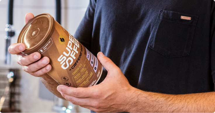 Pessoa segurando uma lata de SuperCoffee Chocolate 380g com as duas mãos, em ambiente interno.