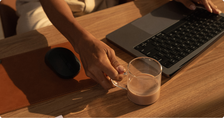 Mão segurando um copo de SuperCoffee ao lado de notebook sobre mesa de madeira.