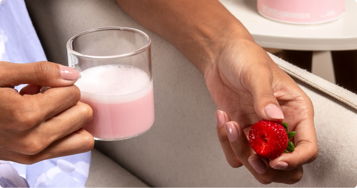 Pessoa segurando uma caneca com bebida rosa cremosa e um morango partido ao meio, sentada em um sofá.
