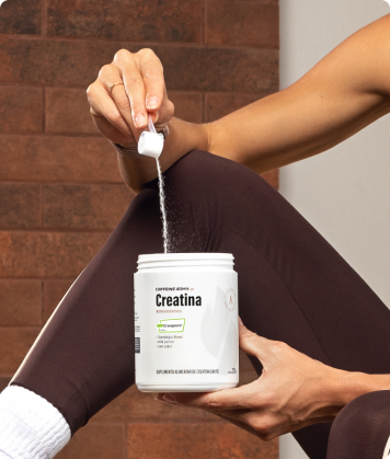 Pessoa adicionando creatina Caffeine Army Labs 250g com colher dosadora sobre o pote, sentada em ambiente interno.