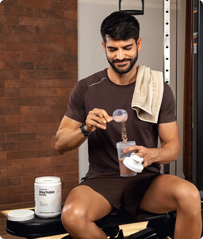 Homem preparando shake com Whey Protein chocolate, adicionando pó ao shaker na academia, com pote aberto ao lado.