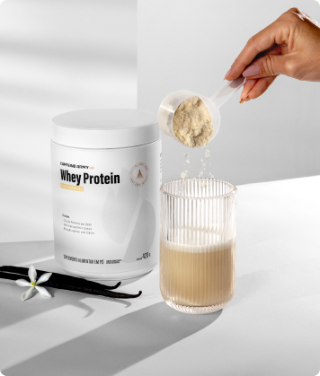 Pessoa adicionando Whey Protein baunilha ao copo com leite ou água, com pote do produto ao fundo e fava de baunilha ao lado.