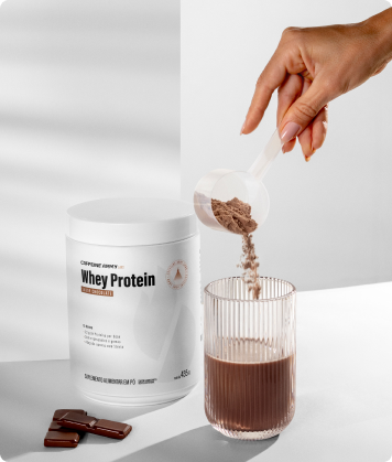Pessoa adicionando Whey Protein chocolate em copo de bebida, com pote do produto e pedaços de chocolate ao lado.